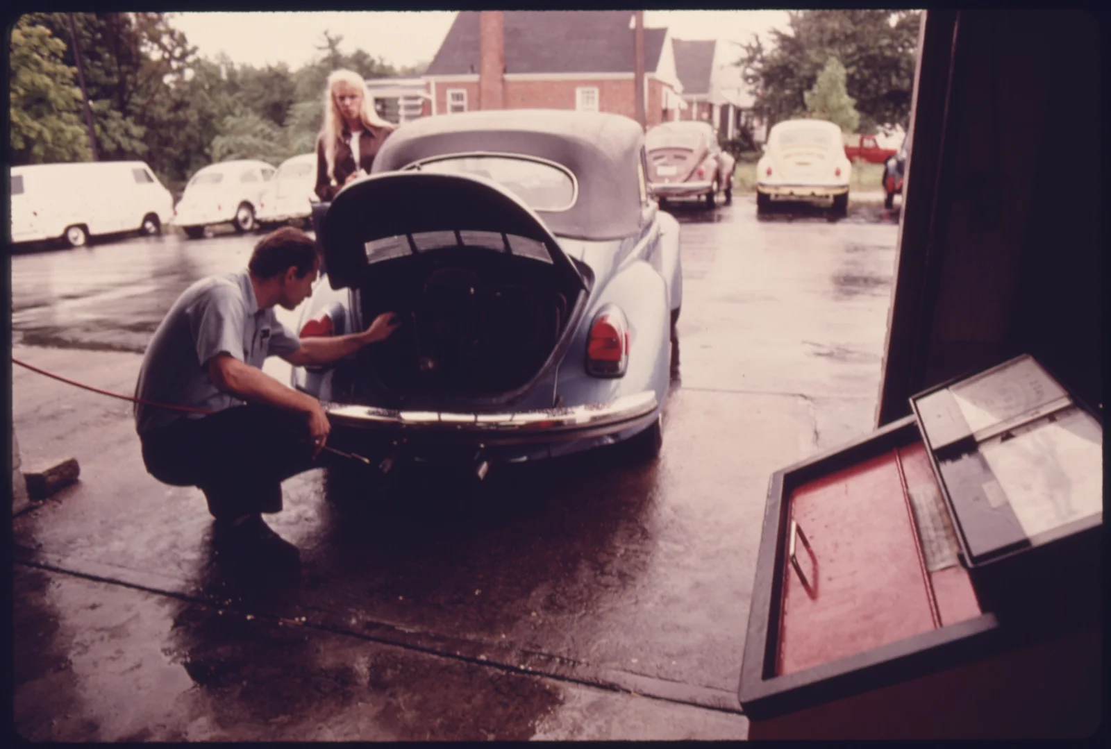 A service station mechanic adjusts the engine while a young woman watches — the classic information gap between expert and customer.